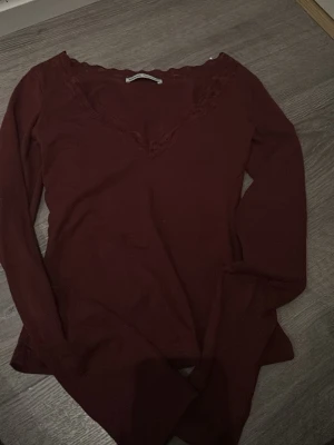 Vinröd långärmad topp från Bershka - Vinröd långärmad topp från Bershka med v-ringning och spetsdetaljer vid halsen. Tunn och mjuk, perfekt att styla med jeans eller kjol. Snygg passform och enkel att matcha med olika accessoarer.