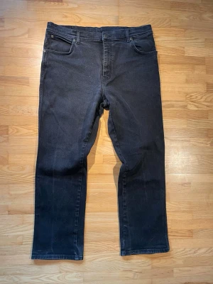 Wrangler jeans svart - Säljer ett par svarta jeans med rak passform och fem fickor. Jeansen har klassiska sömmar och detaljer, samt en diskret patch bak i midjan. Jeansen ger en tidlös vibe. Perfekta för dig som gillar enkel och clean stil.