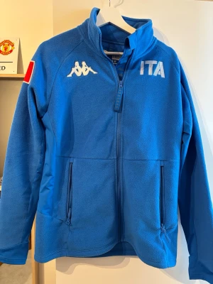 Blå fleecejacka Kappa ITA - Blå fleecejacka från Kappa med broderad ITA-text och logga på bröstet samt italiensk flagga på ärmen. Jackan har hög krage, dragkedja framtill och två sidofickor med dragkedja. Perfekt för kyliga dagar.