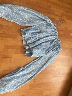 Blå blommig offshoulder blus - Supersöt blå offshoulder blus med vitt småblommigt mönster och långa puffiga ärmar från Gina Tricot. Blusen har resår vid axlar och midja för en snygg passform och är gjord i ett lätt, mjukt tyg som känns luftigt. 