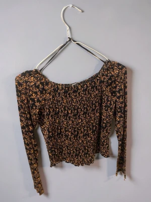 Svart blommig offshoulder topp - Trendig svart offshoulder topp med små blommor i orange och beige. Långärmad modell med smockad kropp och ärmar, vilket ger en tight och stretchig passform. Perfekt för dig som gillar romantisk och feminin stil.