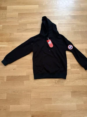 Svart hoodie från Canada Goose - Snygg svart hoodie från Canada Goose med klassisk logga på ärmen och justerbar huva. Hoodien har en känguruficka framtill och ribbade muddar vid ärmslut och nederkant. Perfekt för en chill och stilren look.