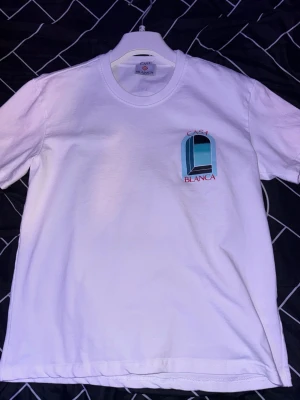 Vit Casa Blanca t-shirt - Säljer en vit t-shirt från Casa Blanca med ett stort grafiskt tryck i blått och svart på ryggen samt mindre tryck på bröstet. T-shirten har rund hals och korta ärmar. Perfekt för dig som gillar stilrena plagg med unik design.
