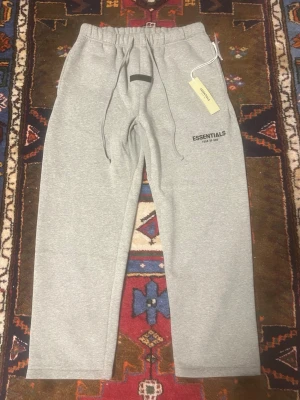 Gråa Essentials mjukisbyxor - Snygga grå mjukisbyxor från Essentials med dragsko i midjan och tryckt logga på ena benet. Byxorna har raka ben och är tillverkade i mjukt material som känns skönt mot huden. Perfekta för en avslappnad stil.