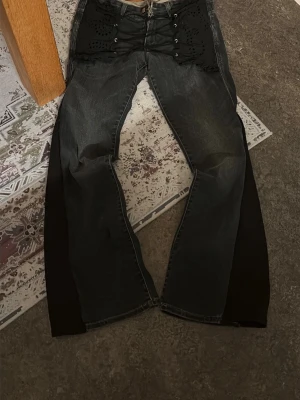 Svarta utsvängda jeans från Kuyichi - Unika svarta jeans från Kuyichi med breda utsvängda ben och coola snörningsdetaljer framtill. Jeansen har svarta tygpaneler på sidorna och dekorativa hålmönster upptill. Perfekta för dig som vill sticka ut med din stil. Sytt bootcut och detaljen. Storlek w29