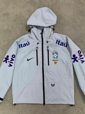 Vit Nike Brasil vindjacka med huva - Snygg vit vindjacka från Nike med Brasil-logga och sponsormärken som Itaú och Vivo i blått. Jackan har huva med dragsko, två bröstfickor med dragkedja och svarta detaljer. Perfekt för dig som gillar sportig stil och vill sticka ut.