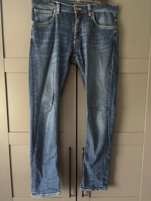 Blå Grim Tim Nudies  - Säljer ett par klassiska blå jeans med raka ben och kontrastsömmar i storlek 33/32(se bild 4). Jeansen har fem fickor, bälteshällor och snygga detaljer på bakfickorna. Materialet är denim i bomull och passformen är avslappnad men ändå stilren. Modellen är Grim Tim och är använda men ändå i väldigt bra skick. Köpta i butik där nypriset låg på 1599kr