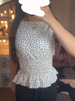 Vit prickig ärmlös topp med volang - Supersöt vit topp med svarta prickar och smockad midja som ger en snygg passform. Toppen är ärmlös och har volangkant nedtill för en extra feminin touch. Perfekt att styla med jeans eller kjol för en trendig look.