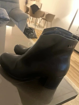 Svarta stövletter med klack från L&M - Snygga svarta stövletter från L&M i slätt skinnliknande material. Modellen har en rundad tå, diskret dragkedja på sidan och stabil blockklack som ger extra höjd. Perfekta för dig som vill ha en stilren och trendig look.