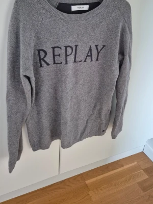 Replay Sweatshirt - Snygg grå stickad tröja från Replay med stor logga framtill. Tröjan har rund halsringning, långa ärmar och en mörkare detalj på ryggen. Perfekt för dig som gillar enkel men stilren design.