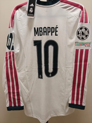 Real Madrid Mbappé spelar version hemma 26/27 - Säljer en långärmad Real Madrid matchtröja med Mbappé och nummer 10 på ryggen. Tröjan är vit med vinröda ränder på ärmarna, svarta detaljer och klubbmärke på bröstet. Tillverkad i funktionsmaterial, perfekt för fotboll eller träning.
