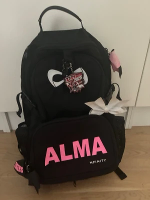Nfinity cheer väska  - Snygg svart ryggsäck från Nfinity med stora rosa bokstäver på fickan framtill och flera fack. Dekorerad med en vit rosett och nyckelringar. Perfekt för dig som vill sticka ut med en personlig touch och mycket plats för dina grejer.Nfinity stor cheerväska. Orginalpris 1700kr och har en defekt på dragkedjan men är i övrigt i bra skick. Texten alma går även att ta bort!