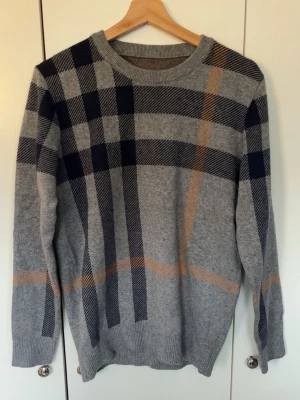 Burberry sweater  - Snygg grå stickad tröja med stora rutiga mönster i mörkblått och beige. Tröjan har rund halsringning, långa ärmar och ribbade muddar vid ärmslut och nederkant. Perfekt för dig som gillar klassisk men trendig stil. Storlek S. Helt ny 