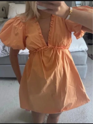 Kort orange klänning med puffärm - Söt kort klänning i orange med puffärmar och djup v-ringning. Klänningen har resår i midjan och volangdetaljer längs ringningen. Perfekt för varma dagar och ger en trendig look.