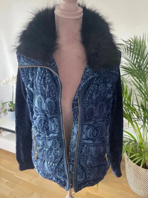 Blå mönstrad dunjacka med fuskpäls - Unik blå dunjacka med detaljerat blommigt mönster i olika blå nyanser. Jackan har en stor krage med svart fuskpäls och guldfärgade dragkedjor. Materialet är mjukt och quiltat, perfekt för kalla vinterdagar. Snygg och trendig design som sticker ut.