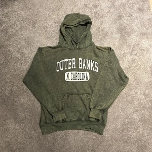 Grön hoodie med Outer Banks-tryck - Cool grön hoodie med vit text 'OUTER BANKS N. CAROLINA' på bröstet. Hoodien har en stor huva och magficka framtill. Materialet är mjukt och har en tvättad vintage-look. Perfekt för en avslappnad streetstyle.