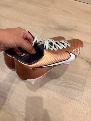 Kopparfärgade Nike fotbollsskor - Snygga fotbollsskor från Nike i kopparfärg med vita detaljer och vita skosnören. Skorna har en låg profil och är designade för bra grepp på planen. Swoosh-logga på sidan och texten 'ACC' tryckt på ovansidan. Perfekta för dig som vill sticka ut på fotbollsplanen.storlek 40,5 