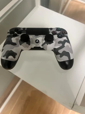 Kamo DualShock 4 handkontroll PS4 - Snygg PlayStation 4 DualShock 4 handkontroll med grå, vit och svart kamouflagemönster på framsidan och svart baksida. Ergonomisk form med klassiska PlayStation-knappar och touchpad. Perfekt för gaming och multiplayer-sessioner. ⭐️Helt felfri, bara att skriva vid minsta lilla⭐️