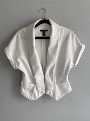 Vit kortärmad kavaj med bred krage - Stilren vit kavaj med korta ärmar och bred sjalkrage. Modellen är kort och har två fickor framtill. Perfekt för att lyfta en outfit med en fräsch och modern touch. Materialet ser ut att vara mjukt och något stretchigt.