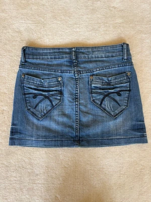 Blå jeanskjol med fickor - 2000- tals lågmidjad jeanskjol i kort modell, med klassiska fickor både fram och bak. Kjolen har bälteshällor, knapp och dragkedja framtill samt dekorativa sömmar på bakfickorna. 37,5 cm midjemått rakt över.