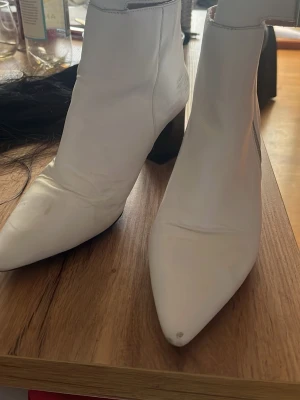 Vita stövletter med klack - Snygga vita stövletter med spetsig tå och bred klack. Skorna har en hög ankel och ett spänne runt skaftet. Tillverkade i slätt material som ser ut som läder. Perfekta för att lyfta vilken outfit som helst.