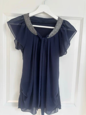 Mörkblå blus med glittrig krage - Det står forever 21 men det e fel märket heter forever. Snygg mörkblå blus i luftig polyester med glittrig, dekorerad krage och knytband framtill. Blusen har korta, fladdriga ärmar och ett elegant fall. Perfekt för dig som vill ha något extra till outfiten.