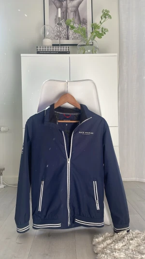 Marinblå vindjacka Race Marine - Snygg marinblå vindjacka från Race Marine med vita dragkedjor och detaljer. Jackan har tryck på bröstet och ärmen, samt två fickor framtill. Tillverkad i lätt och vindtätt material, perfekt för blåsiga dagar. Sportig look med ribbade muddar och krage.