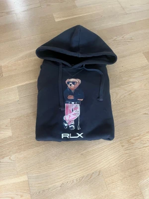 Svart hoodie med björntryck RLX - Svart hoodie med stor huva och dragsnören. På bröstet finns ett färgglatt tryck av en björn med solglasögon och rosa byxor. RLX-logga under trycket. Klassisk känguruficka framtill och ribbade muddar vid ärmar och nederkant.