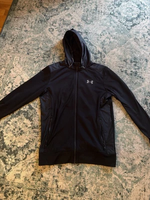 Svart hoodie från Under Armour - Snygg svart hoodie från Under Armour med dragkedja framtill och två fickor med dragkedja. Hoodien har en justerbar huva och logga på bröstet. Tillverkad i ett tekniskt material som andas och känns skönt mot huden.