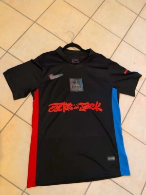 Svart FC Barcelona x Cactus Jack tröja - Säljer en svart FC Barcelona x Cactus Jack matchtröja med röd och blå sidopanel, klubbmärke och Nike-logga på bröstet samt Cactus Jack-tryck framtill. UNHCR-logga på ryggen. Perfekt för dig som gillar unika fotbollströjor.