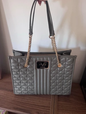 19V69 italia Elenar Shopper Bag - Väska |Handväska | Grön Väska - Stilren grå handväska från Valia med quiltat mönster och guldfärgade kedjedetaljer på handtagen. Väskan har en rektangulär form, dragkedja upptill och flera fack inuti för smart förvaring. Perfekt accessoar för dig som gillar trendiga detaljer.