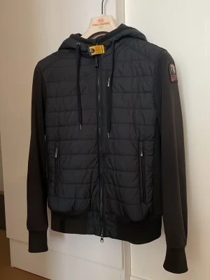 Svart hoodie från Parajumpers - Snygg lätt vår/höst jacka  från Parajumpers med quiltad framsida, dragkedja och två fickor med dragkedja. Har huva med dragsko och ribbade muddar. Patch med logga på ärmen och gul detalj vid halsen. Perfekt för en cool och avslappnad stil.
