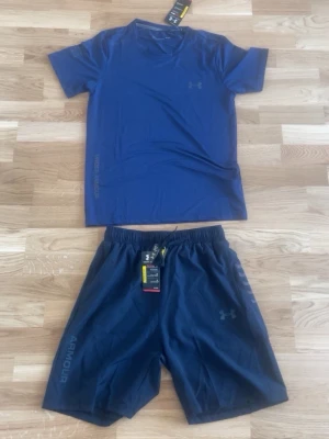Blått träningsset från Under Armour - Snyggt blått träningsset från Under Armour med t-shirt och shorts. Setet är gjort i ett lätt och snabbtorkande material, perfekt för träning. T-shirten har rund hals och kort ärm, shortsen har resår i midjan och logga på benet.