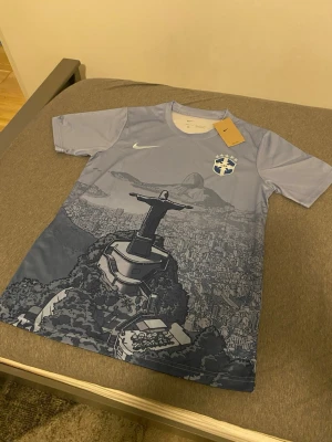 Nike Brasil fotbollströja blå - Snygg ljusblå fotbollströja från Nike med Brasilien-emblem och motiv av Kristusstatyn i Rio de Janeiro. Tröjan har rund hals och korta ärmar, perfekt för träning eller match. Materialet är lätt och andas bra.