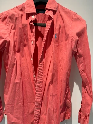 Korallfärgad skjorta från Polo Ralph Lauren - Säljer en snygg korallfärgad skjorta från Polo Ralph Lauren med klassisk krage och blå broderad logga på bröstet. Skjortan har långa ärmar, knappar framtill och är tillverkad i mjuk bomull. Perfekt för dig som vill ha en stilren och färgglad look.
