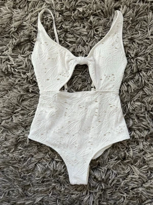 Vit baddräkt med broderade detaljer - Snygg vit baddräkt med broderade blommönster och dekorativ knytning framtill. Baddräkten har smala axelband och ett litet hål under bysten. Aldrig använda!!