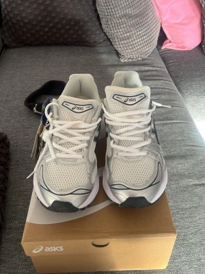 Vita Asics sneakers med meshdetaljer - Snygga vita sneakers från Asics med meshpaneler för extra ventilation och detaljer i silver och svart. Klassisk låg modell med vita skosnören och bekväm vadderad plös. Perfekta för dig som gillar sportig och clean stil.