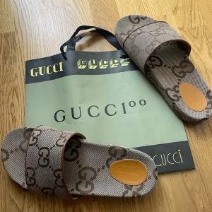 Sandaler  - Använda en gång, påsen ingår, inte äkta            Säljer ett par snygga sandaler i beige och brunt med det klassiska GG-monogrammet över hela skon. Öppen tå och bred rem över foten. Tillverkade i canvasliknande material med bruna detaljer. Perfekta för en trendig och avslappnad stil.