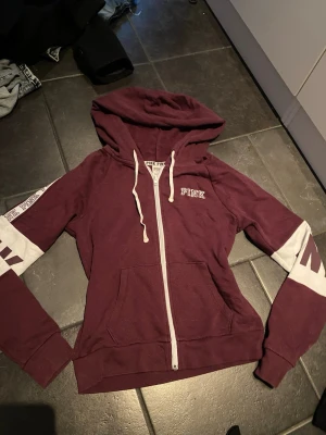Vinröd hoodie med dragkedja från PINK - Snygg vinröd hoodie från PINK med vit dragkedja och huva. Ärmarna har vita partier med vinröda detaljer och texten 'PINK' längs axeln. Två fickor framtill och ribbade muddar. Perfekt för en avslappnad och trendig look.