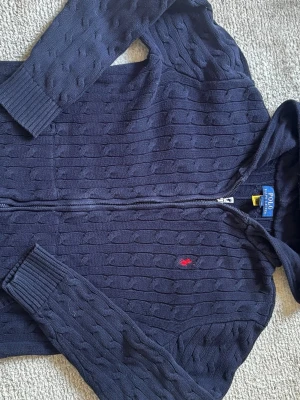 Marinblå kabelstickad hoodie Polo - Snygg marinblå kabelstickad hoodie från Polo Ralph Lauren med dragkedja och huva. Tröjan har ribbade muddar och det klassiska röda Polo-märket broderat på bröstet. Perfekt för en avslappnad men stilren look.