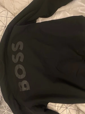 Svart sweatshirt med BOSS-tryck - Säljer en svart sweatshirt med stort BOSS-tryck på framsidan. Tröjan har rund halsringning och långa ärmar. Materialet känns mjukt och bekvämt, perfekt för en avslappnad stil. Passar dig som gillar enkel men snygg design.