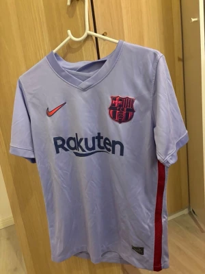 Ljuslila FC Barcelona matchtröja Nike - Säljer en ljuslila FC Barcelona matchtröja från Nike med klubbmärke och Rakuten-tryck på bröstet. Tröjan har röda och blå detaljer, korta ärmar och V-ringning. Tillverkad i lätt och ventilerande material, perfekt för fotboll eller träning.