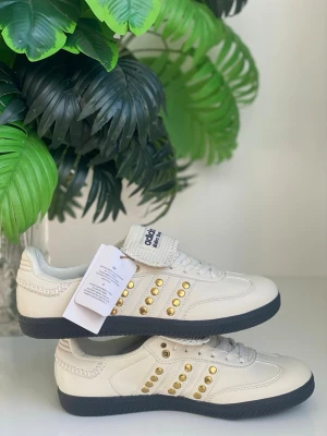 Adidas sneakers med gulddetaljer - Snygga Adidas sneakers i beige läder med klassiska tre ränder på sidan, dekorerade med guldfärgade nitar. Skorna har svart sula och vit ovandel med diskreta sömmar. Perfekta för dig som vill sticka ut med stilrena detaljer.