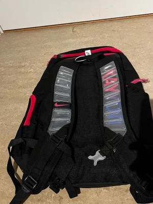 Svart ryggsäck med rosa detaljer - Snygg svart ryggsäck med rosa dragkedjor och detaljer. Axelremmarna har genomskinliga plastpaneler och texten 'KAYY' i rosa. Ryggsäcken har flera fack och justerbara remmar, perfekt för skolan eller träning.