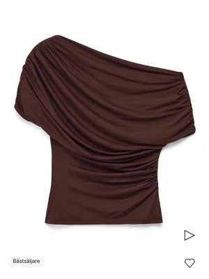 Brun oneshoulder topp med drapering - Snygg brun topp med oneshoulder-design och draperade detaljer. Toppen har en tight passform och är tillverkad i ett mjukt, stretchigt material som sitter skönt på kroppen. Perfekt för dig som vill sticka ut med en unik siluett. Aldrig använd med prislappen kvar 
