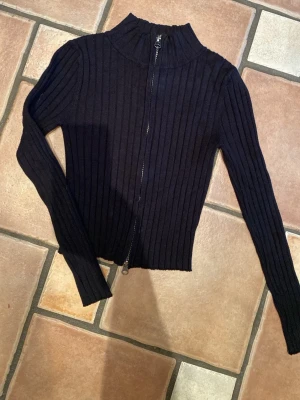 Svart ribbad ziptröja från H&M - Snygg svart ribbstickad tröja från H&M med dragkedja framtill och hög krage. Tröjan har långa ärmar och är figurnära i modellen, perfekt att matcha med jeans eller kjol. Materialet är mjukt och stretchigt för extra komfort.