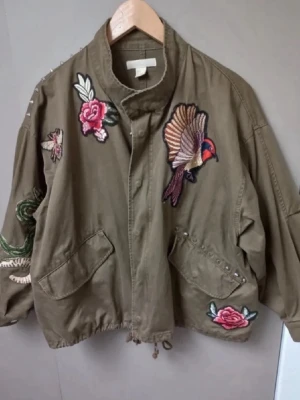 Viral jacka  - KAN INTE POSTA FÖREN 11 SEPTEMBER!! Säljer en olivgrön vindjacka med coola broderier av fågel, rosor och fjäril. Jackan har två stora fickor framtill, dragsko i nederkant och dekorativa nitar vid ena fickan. Oversized passform och ståkrage ger en avslappnad look.