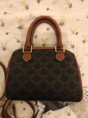 Brun handväska med monogram - Snygg handväska från  i mörkbrunt canvasmaterial med klassiskt monogrammönster och detaljer i ljusbrunt läder (ska vara läder enligt butiken jag köpte från men reserverar mig för eventuellt fin vegansk läder). Väskan har guldfärgade metalldetaljer, dubbla handtag och avtagbar axelrem. Perfekt för dig som gillar lyxig och tidlös design. Ingår två axelband och en dustbag. Använd ett par gånger. Storlek cirka 20cm. Priset är mycket bra och är inte förhandlingbart.