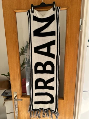 Svartvit halsduk med text - Säljer en lång, stickad halsduk i svart och vitt med stora bokstäver som bildar ordet 'URBAN' på båda sidor. Halsduken har fransar i ändarna och en cool, grafisk design som sticker ut. Perfekt accessoar för att lyfta din outfit.