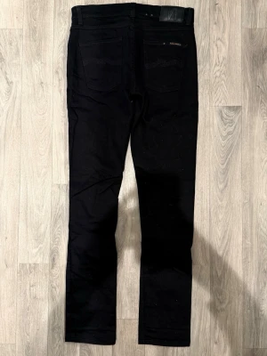 Nudie Jeans Lean Dean Svarta W30/L34 - Slim Tapered Fit - Nudie Jeans Lean Dean ”Dark Ever Black” svarta jeans med slim tapered-passform, normalhög midja och avsmalnande ben, storlek W30/L34, midja 39 cm, ytterbenslängd 106 cm, benöppning 16 cm, modellen är 185cm & väger 65kg, i fint skick.